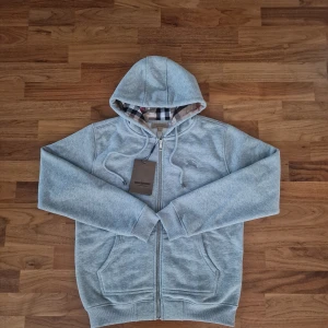 Burberry Hoodie - Snygg grå hoodie från Burberry med klassiskt rutigt foder i huvan. Tröjan har hel dragkedja, två fickor fram och justerbar huva med snörning. Tillverkad i mjuk bomullsmix för extra komfort. Perfekt för en chill och stilren look.