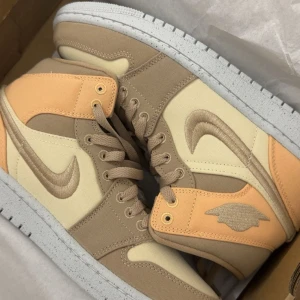 Nike Jordan 1 i beige och persika - Säljer ett par Nike Jordan 1 sneakers i snygga beige, persika och krämfärgade toner. Skorna har broderad swoosh, snörning och vit sula. Tillverkade i canvasmaterial med klassisk high-top design. Perfekta för dig som vill sticka ut med stilrena färger.