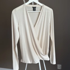Beige/ vit omlottopp från Lindex XS - Säljer en stilren beige omlottopp från Lindex i storlek XS. Toppen har lång ärm, v-ringning och ett snyggt omlottfall framtill. Tillverkad i ett mjukt och lätt material som ger en elegant känsla.🌸liten fläck vid kragen  smink 