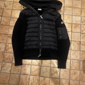 Svart Moncler pufferjacka med huva - Svart pufferjacka från Moncler med stickade ärmar och quiltad front. Jackan har huva, dragkedja framtill och Moncler-logga på ärmen. Materialmix av dun, polyester och stickad ull ger både värme och stil. Perfekt för kyliga dagar. Vet ej om äkta och scannen funkar inte 