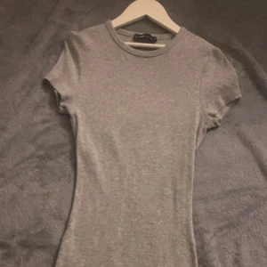 Grå basic klänning i bomull - Enkel och stilren grå t-shirt med rund hals och korta ärmar. Modellen är figurnära och passar perfekt till jeans eller kjol. Gjord i mjuk bomull för maximal komfort.