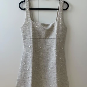 Beige pärlprydd kort klänning H&M - Superfin beige kort klänning från H&M med pärldetaljer framtill. Klänningen har breda axelband, fyrkantig ringning och är tillverkad i ett glittrigt, strukturerat material som ger en lyxig känsla. Perfekt för dig som vill sticka ut med en elegant och trendig look.