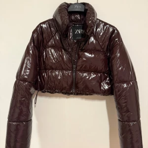 Croppad brun pufferjacka från Zara - Säljer en croppad, glansig pufferjacka från Zara i mörkbrunt/vinrött. Jackan har hög krage, lång ärm och det går att justera midjan (se sista bild). Storlek xs. 
