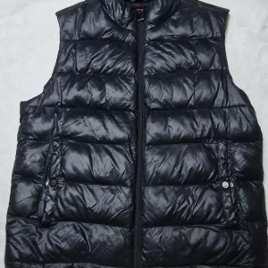 Svart dunväst från Hugo - Svart dunväst från Hugo med glansig finish och diskret logga på bröstet och ryggen. Västen har ståkrage, två fickor med knappdetalj och dragkedja framtill. Perfekt för lager-på-lager och en clean streetstyle-look.