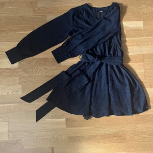 Svart prickig omlottklänning Bik Bok - Säljer en svart kort omlottklänning från Bik Bok med prickigt mönster. Klänningen har långa ärmar med breda manschetter och knappar, v-ringad hals och ett knytband i midjan som ger en snygg siluett. Perfekt för dig som gillar stilrena detaljer och prickigt tyg.