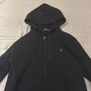 Svart hoodie från Polo Ralph Lauren - Svart hoodie från Polo Ralph Lauren med dragkedja och huva. Klassisk design med broderad logga på bröstet och snörning i huvan. Perfekt för en chill och stilren look. Materialet är mjukt och skönt, troligtvis bomullsmix.