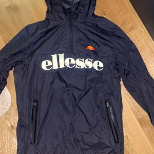Svart vindjacka från Ellesse - Svart vindjacka från Ellesse med stor logga framtill, huva och två dragkedjeförsedda fickor. Jackan har half zip och är tillverkad i lätt polyester, perfekt för blåsiga dagar. Snygg och sportig stil som passar till streetwear.