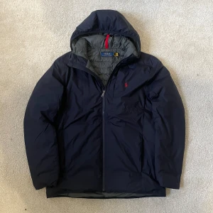 Marinblå pufferjacka från Polo Ralph Lauren | M - Svart pufferjacka från Polo Ralph Lauren med huva och dragkedja framtill. Jackan har en diskret röd logga broderad på bröstet och grått foder på insidan. Perfekt för kyliga dagar och riktigt snygg streetstil.