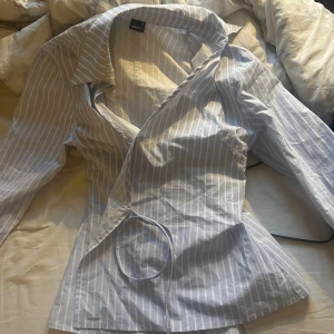 Randig omlottblus från Gina Tricot - Snygg ljusblå och vit randig omlottblus från Gina Tricot. Skjortan har långa ärmar, klassisk krage och knytband i midjan för en schysst passform. Perfekt för dig som gillar stilrena och fräscha looks.