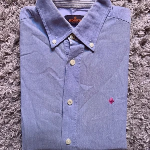 Blå skjorta från Morris - Snygg blå skjorta från Morris med klassisk krage och vita knappar. Skjortan har en liten rosa broderad logga på bröstet och är tillverkad i mjuk bomull. Perfekt för dig som gillar stilrena och tidlösa plagg.