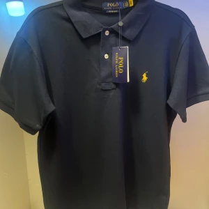 Royal blå piké Polo Ralph Lauren - Klassisk blå pikétröja från Polo Ralph Lauren i bomull med gul broderad logga på bröstet. Tröjan har korta ärmar, krage och knappar framtill. Slim fit-modell som sitter snyggt och stilrent. Perfekt för en clean och tidlös look.