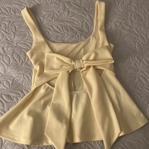 Gul linnetopp med stor rosett - Supersöt ljusgul linnetopp med breda axelband och stor rosett baktill . Toppen har en lätt utsvängd form och en snygg cut-out under rosetten. Perfekt för dig som vill sticka ut med en feminin och trendig look. Priset går att diskutera 