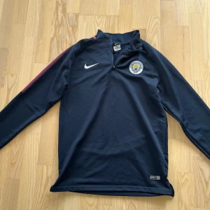 Manchester City långärmad träningströja Nike - Mörkblå långärmad Manchester City träningströja från Nike med halv dragkedja och klubbmärke på bröstet. Tröjan har en röd detalj längs ena ärmen och är tillverkad i ett mjukt, sportigt material som passar perfekt för fotbollsträning.
