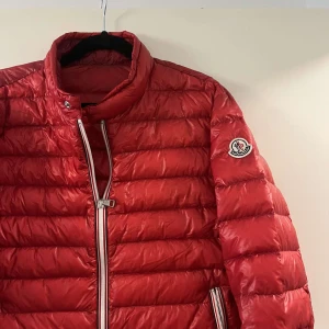 Röd dunjacka från Moncler - Säljer en röd dunjacka från Moncler med klassisk quiltad design och hög krage. Jackan har vita detaljer vid dragkedjorna och Moncler-logga på ärmen. Perfekt för kyliga dagar och riktigt snygg till streetwear-stilen.