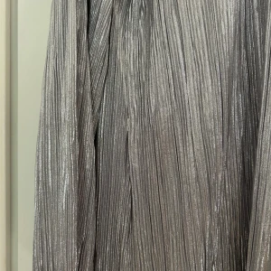 Glittrig silverklänning med volanger - Säljer en glittrig silverfärgad klänning med plisserat tyg och lång ärm. Klänningen har snygga volangdetaljer nedtill som ger extra rörelse och attityd. Perfekt för dig som vill sticka ut med metallic och coola detaljer.
