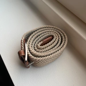 Beige flätat bälte med skinn-detaljer - Snyggt beige flätat bälte med bruna skinn-detaljer och silverfärgat spänne. Bältet har en stilren och modern look med twistad textur, perfekt för att lyfta vilken outfit som helst.