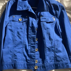 Blå jeansjacka från Ted Nicol - Snygg blå jeansjacka från Ted Nicol med klassisk krage, guldiga knappar och två bröstfickor med lock. Jackan har en rak passform och är tillverkad i bomull, perfekt för dig som vill ha en stilren och tidlös look.