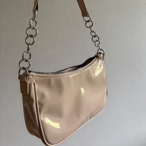Beige axelväska med kedjedetaljer - Snygg beige axelväska i glansigt syntetmaterial med silvriga kedjedetaljer på axelremmen. Väskan har en dragkedja upptill och ett rymligt innerfack. 