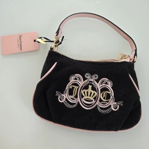Svart axelväska från Juicy Couture - Söt svart axelväska från Juicy Couture i mjuk frotté med ljusrosa detaljer och broderat JC-emblem med krona framtill. Baksidan har ett hjärtformat rosa läderpatch med guldfärgad hjärtdetalj. Väskan har guldfärgad dragkedja och matchande axelrem.