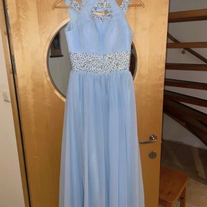 Ljusblå långklänning med strassdetaljer - Superfin ljusblå långklänning med plisserad överdel och bred midja dekorerad med glittrande strass. Klänningen har ärmlös design med snygga axelband och ett flowy, svepande kjolparti. Perfekt för bal eller festliga tillfällen! Mått runt midjan: 79cm Min längd från axel och ner: 140 cm Mått över bysten: 94cm Står ingen storlek men sitter som en XXS/XS