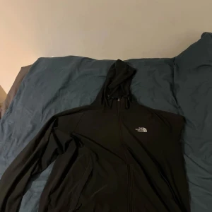 Svart vindjacka från The North Face - Svart vindjacka från The North Face med huva och dragkedja framtill. Jackan har en clean look med logga på bröstet och är tillverkad i ett lätt och smidigt material. Perfekt för blåsiga dagar och enkel att matcha med det mesta.