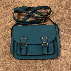 Blå axelväska med spännen - Snygg blå axelväska i syntetmaterial med justerbar rem och klassiska spännen framtill i guldton. Väskan har flera fack, inklusive ett med dragkedja, och en praktisk ficka på baksidan. Perfekt för att bära dina viktigaste grejer med stil.