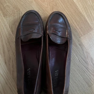 Bruna loafers från Prada i skinn - Klassiska bruna loafers från Prada i äkta skinn. Snygg penny loafer-design med rund tå och låg klack. Perfekta för dig som gillar tidlös och stilren look. Mörkbrun färg med diskreta sömmar och detaljer.