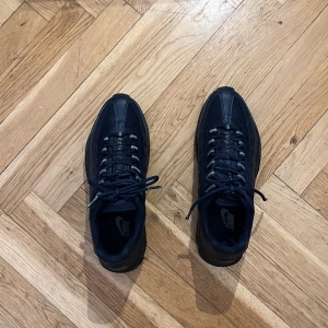 Nike Air Max 95 svarta sneakers - Säljer ett par svarta Nike Air Max 95 sneakers med klassisk chunky sula och synliga Air-enheter. Skorna har snörning, rund tå och är tillverkade i en mix av syntet och textil. Perfekta för dig som gillar sportig och stilren look. Har slitningar på skosnörena