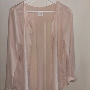 Ljusrosa blus från Vila i storlek S - Säljer en ljusrosa blus från Vila med lång ärm och knappar vid ärmslutet. Blusen är tunn och har en snygg knyt-detalj framtill. Perfekt för dig som gillar en stilren och feminin look.
