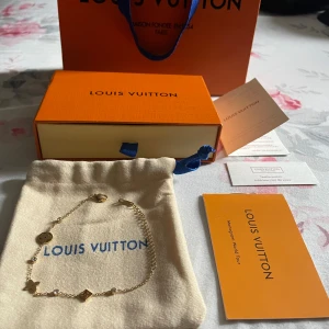 Louis Vuitton guldarmband med kristaller - Exklusivt armband från Louis Vuitton i guldtonad metall med ikoniska LV-symboler och gnistrande kristaller. Kedjan har flera detaljer, bland annat en svart blomma och geometriska former. Levereras med originalpåse, ask och certifikat.
