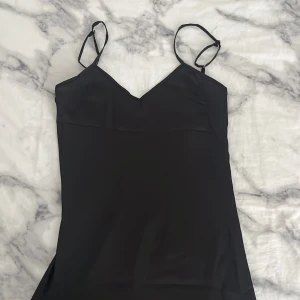 Svart satin långklänning  - Supersnygg svart långklänning med smala axelband och v-ringad front. Klänningen har en öppen rygg med spetsdetaljer och är gjord i ett glansigt satinliknande material som faller mjukt. Perfekt för dig som gillar stilren och elegant look.