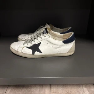Golden Goose Superstar vita sneakers - Golden Goose Superstar sneakers i vitt skinn med svart stjärndetalj på sidan och mörkblå häl. Klassisk låg modell med grå snörning och platt sula. Märkeslogga på plösen och insidan. Perfekta för dig som gillar en cool och avslappnad stil.