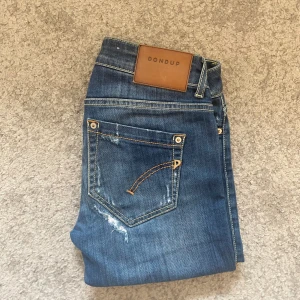 Blå Dondup Monroe jeans med slitningar - Säljer ett par blå Dondup Monroe jeans med snygga slitningar och kontrastsömmar. Klassisk femficksmodell med läderpatch bak i midjan och coola detaljer på bakfickorna. 