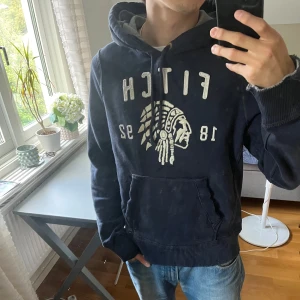 Mörkblå hoodie från Abercrombie & Fitch - Säljer en mörkblå hoodie från Abercrombie & Fitch med vit broderad logga och motiv framtill. Tröjan har några fläckar men är ändå riktigt snygg.