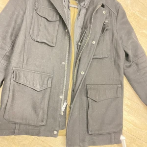 Svart parkajacka med stora fickor - Svart parkajacka med fyra stora fickor framtill och dragkedja samt knappar. Jackan har en klassisk rak passform och är fodrad för extra värme. Perfekt för kyliga dagar och har en stilren look som funkar till det mesta.