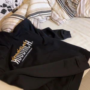 Svart hoodie från Hoodrich med tryck - Svart hoodie från Hoodrich med vit och gul broderad logga framtill och tryck på ryggen. Klassisk huva, lång ärm och känguruficka. Perfekt streetwear-vibe och riktigt snygg till jeans eller joggers.