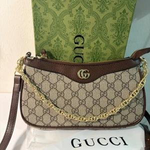 Ophidia Brun i klassiskt GG - Snygg Ophidia i klassiskt GG Supreme-mönster. Väskan är beige och brun med detaljer i brunt skinn, guldfärgad kedja och dragkedja. Ikonisk GG-logga framtill och innerfack i ljust tyg. Perfekt för dig som vill ha en lyxig och trendig accessoar. Skickas snabbt med kartong och dustbag. 