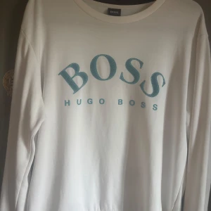 Vit sweatshirt från Hugo Boss - Vit långärmad sweatshirt från Hugo Boss med stort blått BOSS-tryck på bröstet. Tröjan har rund halsringning och är gjord i mjuk bomull, perfekt för en clean och stilren look. Pris är diskuterbart 