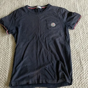  Äkta Mörkblå Moncler t-shirt med ficka - Mörkblå t-shirt från Moncler med korta ärmar och coola rödvita detaljer vid ärmslut och hals. Bröstficka med Moncler-logga ger extra stilpoäng. Perfekt för dig som gillar klassisk design med en twist.
