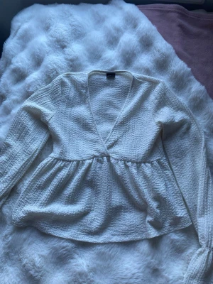 Vit omlottopp med v-ringning - Säljer en vit omlottopp med v-ringning och lång ärm. Toppen har en lätt strukturerad yta och ett löst fall nedtill som ger en avslappnad siluett. Perfekt att styla med jeans eller kjol för en trendig look.
