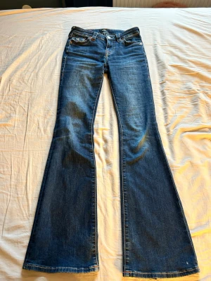 Blå bootcut jeans med fickdetaljer - Säljer ett par blå bootcut jeans med snygga fickdetaljer bak och klassisk femficksdesign. Jeansen har låg midja och är tillverkade i ett slitstarkt denimtyg. Perfekta för dig som gillar en retroinspirerad look med utsvängda ben.