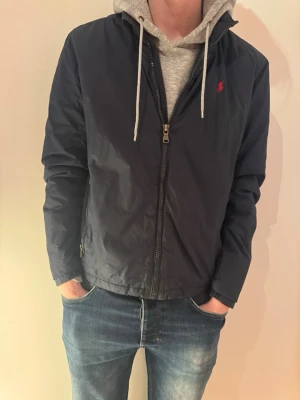 Mörkblå vindjacka från Ralph Lauren - Snygg mörkblå vindjacka från Ralph Lauren med dragkedja framtill och diskret röd logga på bröstet. Jackan har lång ärm och klassisk passform, perfekt för vår och höst. Tillverkad i lätt polyester som står emot vind.
