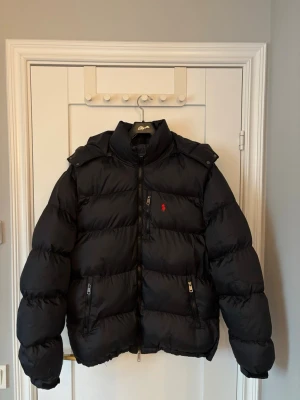 Svart pufferjacka från Polo Ralph Lauren - Snygg svart pufferjacka från Polo Ralph Lauren med röd logga på bröstet. Jackan har hög krage, dragkedja framtill och flera praktiska fickor med dragkedja. Perfekt för Svenskt väder. Säljer då den inte används längre, skick 10/10
