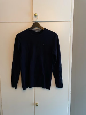Mörkblå stickad tröja från J.Lindeberg - Snygg mörkblå stickad tröja från J.Lindeberg med rund halsringning och diskret logga på bröstet. Sparsamt använd. Står att den är M men skulle säga att den passar mer som S. Tveka inte att fråga om du undrar något.