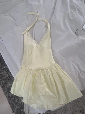 Ljusgul kort klänning med knyt - Söt ljusgul kort klänning med v-ringning och knyt i nacken. Klänningen har en volangig kjol och soprent uttryck, perfekt för varma dagar. Tillverkad i ett lätt tyg som känns luftigt och bekvämt.