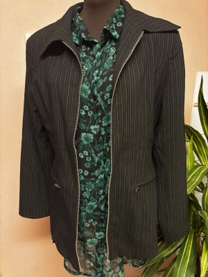 Svart randig kavaj med dragkedja - Snygg svart kavaj med tunna vita ränder och dragkedja framtill. Jackan har en klassisk krage, lång ärm och fickor med dragkedja. Axelkuddar. Passar perfekt till en trendig och stilren look.