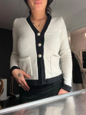 Vit cardigan med svarta detaljer - Stilren vit cardigan med svarta kanter och dekorativa pärlknappar. Tröjan har två fickor framtill och är långärmad. Perfekt för dig som gillar klassisk och chic stil.