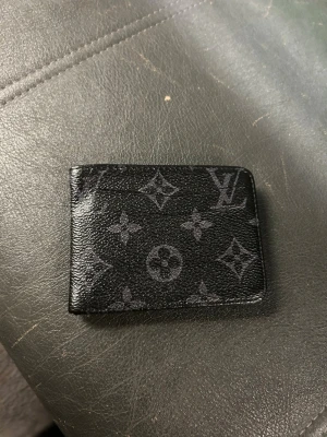 Svart snygg plånbok - Snygg svart plånbok med klassiskt monogrammönster i svart. Plånboken är tillverkad i skinn och har flera fack för kort. Perfekt accessoar för dig som gillar stilrena detaljer och lyxig känsla.