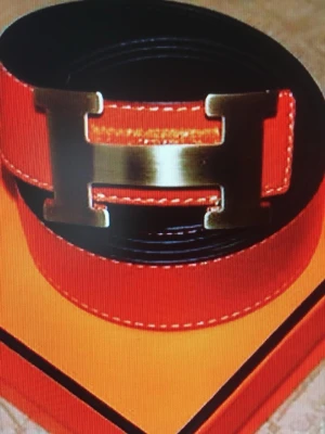 H  orange läderbälte med H-spänne - Snyggt orange bälte från i läder med ikoniskt H-format metallspänne i guldton. Bältet har vita kontrastsömmar och klassisk design som ger en exklusiv känsla. Perfekt accessoar för att lyfta vilken outfit som helst.