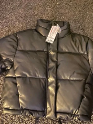 Svart pufferjacket - Snygg svart pufferjacka med croppad passform och hög krage. Jackan har quiltad design och dragsko i nederkant för extra stilpoäng. Tillverkad i glansigt syntetmaterial som ger en modern look. Perfekt för dig som vill ha en trendig och varm jacka.